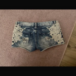 Blå jeansshorts med spets och pärlor - Snygga blå jeansshorts med ljus tvätt, med vit spets och små pärlor på sidorna. Shortsen har slitna detaljer vilket verkligen är jättesnyggt. Säljer pågrund av att de är för små för mig. De är ganska små i storleken men passar xs-s storlek.  Midjemått 37cm❤️Dålig bild men där låren sitter är måtten 24-24 cm ungefär❤️