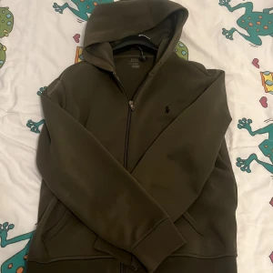 Olivgrön hoodie från Polo Ralph Lauren - Snygg olivgrön hoodie från Polo Ralph Lauren med dragkedja och klassisk broderad logga på bröstet. Tröjan har huva, långa ärmar och praktiska fickor framtill. Perfekt för en chill och stilren look funkar både för M|L.