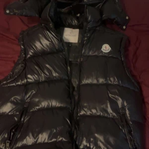 Svart väst moncler  - Snygg svart dunväst från Moncler med glansig finish och klassisk logga på bröstet. Västen har hög krage, dragkedja framtill och två fickor med dragkedja. Insidan har en cool serietidningsdetalj. Perfekt för lager-på-lager-stil.