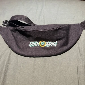 Svart magväska STHLM SÖDRA brodyr - Svart magväska i slitstark polyester med broderad STHLM SÖDRA-logga i grönt, gult och blått framtill. Väskan har justerbart band med spänne och dragkedja upptill. Perfekt för att bära det viktigaste på stan eller till matchen.