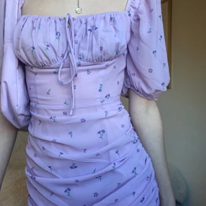 Ljuslila blommig kortklänning från Adoore - Supersöt ljuslila kortklänning med små blommor i lila och grönt. Klänningen har puffiga korta ärmar, rynkade detaljer och knytning vid bysten. Sitter figurnära och har en romantisk vibe, perfekt för vår och sommar. Säljer då den inte kommer till användning, endast använda fåtal gånger! Skriver för fler bilder eller frågor!