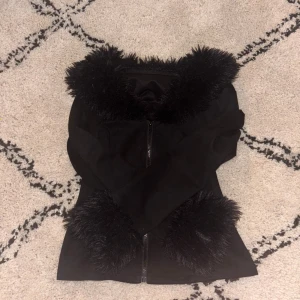 Svart pälskofta med dragkedja - Säljer en svart pälskofta med fluffiga detaljer vid krage och ärmslut. Jackan har en snygg figurnära passform och stängs med dragkedja framtill. Materialet är mjukt och perfekt för dig som vill sticka ut med en cool look.