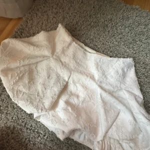 Vit kort kjol med broderade detaljer - Supersöt vit kort kjol med broderade blommor och volangkant. Kjolen har en lätt utsvängd form och ger en romantisk vibe. Perfekt för dig som gillar ljusa och feminina plagg med det lilla extra.