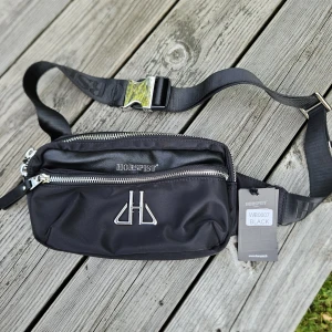 Svart magväska från Horspist - Snygg svart magväska från Horspist med silverfärgade dragkedjor och logga framtill. Väskan har justerbart band med spänne och flera fack för smidig organisering. Tillverkad i slitstark polyester och har en clean, modern look.