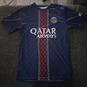 PSG matchtröja Qatar Airways - Snygg PSG fotbollströja i mörkblått med röd och vit rand längs mitten, Qatar Airways-tryck på framsidan och spelarnamn D. Doué samt nummer 14 på ryggen. Tröjan har korta ärmar och är tillverkad i ett lätt, ventilerande material perfekt för fotboll.