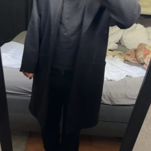Svart lång kappa i ullmix - Säljer en svart, stilren och lång kappa med rak passform. Jackan har stora fickor framtill och är perfekt för dig som gillar en clean och minimalistisk look. Materialet är en skön ullmix som håller dig varm under kyliga dagar.