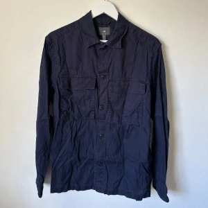 Mörkblå overshirt från H&M i linne - Snygg mörkblå overshirt från H&M i regular fit. Skjortan har två stora bröstfickor med lock, knäppning framtill och är tillverkad i en skön mix av linne och bomull. Perfekt att bära öppen eller stängd för en avslappnad stil. B317