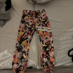 Färgglada leggings från Stronger, S - Säljer ett par leggings från Stronger i storlek small med ett galet fruktigt mönster i rosa, gult, grönt och orange. De är tillverkade i stretchigt material (85% polyester, 15% elastan) och har vita ränder på ena benet. 