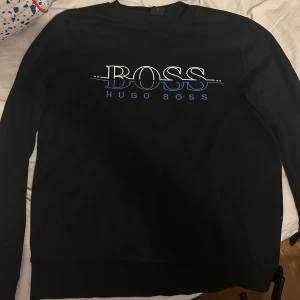 Svart Hugo Boss sweatshirt med logga - Svart sweatshirt från Hugo Boss med stor vit och blå BOSS-logga på bröstet. Tröjan har rund halsringning och långa ärmar. Perfekt för dig som gillar stilrena och klassiska plagg med en modern touch. (Börjar bli för liten)