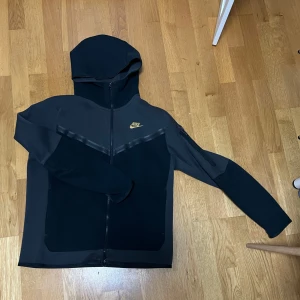 Svart grå nike tech med guld logga - Nike hoodie i svart med dragkedja och huva. Snyggt guldigt Nike-märke på bröstet. Lite gammal men känns inte så.