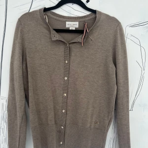 Beige kofta från Holly & Whyte - Snygg beige kofta från Holly & Whyte med rund hals och långa ärmar. Knäppning framtill med guldiga knappar. Mjuk och tunn stickad kvalitet som passar perfekt till lager-på-lager. Enkel och stilren design.