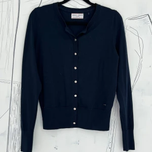 Mörkblå kofta från Holly & Whyte - Snygg mörkblå kofta från Holly & Whyte med rund hals och guldiga knappar framtill. Modellen är klassisk med långärmade och ribbade muddar. Perfekt att slänga över en t-shirt eller linne för en chill look. Materialet är mjuk bomull.