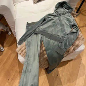 Grön velour hoodie och byxor från Juicy Couture - Mjuk och lyxig gröna hoodie och byxa från Juicy Couture i velour, snörning på byxan och dragkedja framtill på tröjan. Tröjan har långa ärmar, ribbade muddar och en broderad logga på bröstet. Perfekt för en chill och trendig look.