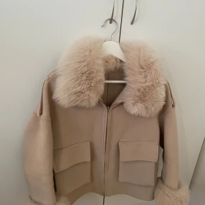 Beige ulljacka med fuskpälsdetaljer - Säljer en beige jacka i ull med stora fickor framtill och dragkedja. Kragen och ärmsluten har fluffiga fuskpälsdetaljer som ger en lyxig vibe. Jackan har en loose passform och är perfekt för höst och vinter. Superfin till både jeans och kjol!