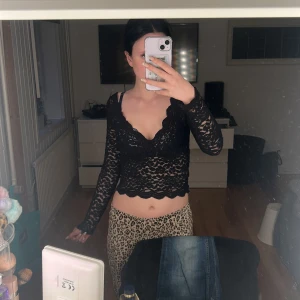 Svart spetsig croppad blus - Säljer en svart långärmad blus i spets med v-ringad hals och vågig kant. Toppen är croppad och har transparent spetsmaterial som ger en cool och edgy vibe. Perfekt för dig som gillar att sticka ut med detaljerad design.