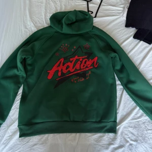 Grön hoodie från Romwe med tryck - Grön hoodie från Romwe i storlek L med rött 'Action'-tryck och stjärnor på både fram- och baksida. Tröjan har huva med svarta snören och en stor magficka. Perfekt för dig som gillar streetstyle och vill sticka ut med färg. Använt totalt 3 gånger 