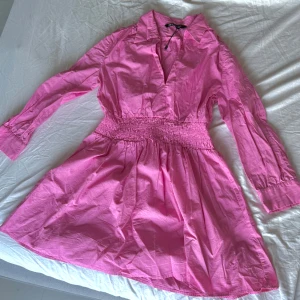 Rosa kortklänning från Zara - Supersöt rosa kortklänning från Zara med långa ärmar, markerad midja med smock och klassisk krage. Klänningen har en lätt v-ringning och är gjord i ett tunt, skönt bomullstyg. Perfekt för dig som vill sticka ut med färg!