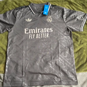 Grå Real Madrid fotbollströja Adidas XL - Snygg grå Real Madrid fotbollströja från Adidas i storlek XL. Tröjan har korta ärmar, krage med knapp och diskret mönster över hela tyget. Broderat klubbmärke och Adidas-logga på bröstet, samt 'Emirates Fly Better' tryckt framtill. Tillverkad i lätt och ventilerande material.