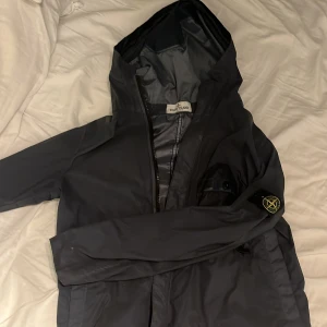 Grå vindjacka från Stone Island Junior - Grå vindjacka från Stone Island Junior med huva och klassisk logotyp-patch på ärmen. Jackan har dragkedja framtill och är tillverkad i lätt polyester som ger en clean och sportig look. Perfekt för dig som gillar streetwear och vill ha något stilrent.