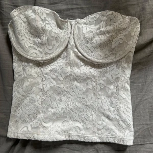 Vit spetskorsett med blommönster - Supersnygg vit korsett med blommig spets och vadderade kupor. Toppen är axelbandslös och har en bandeauformad siluett. Baksidan har smock för extra komfort och passform. Perfekt att styla med jeans eller kjol för en trendig look.