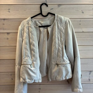Beige quiltad jacka från Amisu - Snygg beige jacka från Amisu med quiltade detaljer framtill och på axlarna. Jackan har dragkedjor både framtill och vid ärmsluten samt två fickor med dragkedja. Materialet känns mjukt och har en mockaliknande yta. 