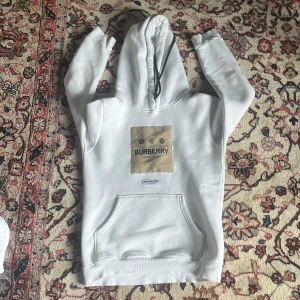 Vit Burberry hoodie med logga - Snygg vit hoodie från Burberry med stor beige logga framtill och klassisk känguruficka. Tröjan har huva med snörning och långa ärmar. Perfekt för dig som vill ha en clean och stilren look med exklusiv känsla.