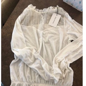 Vit blus med spets  - Supersöt vit blus med lång ärm och elastisk midja Överdelen har fina spetsdetaljer och lätt rynkad halsringning. Perfekt för dig som gillar romantisk och feminin stil.