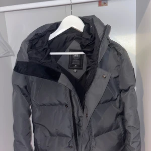 Grå dunjacka från Canada Goose - Säljer en grå dunjacka från Canada Goose med svart logotyp på ärmen. Jackan har huva med dragsko, dragkedja och tryckknappar framtill. Perfekt för kalla dagar och riktigt snygg streetstil. Fodrad och med klassisk pufferlook.