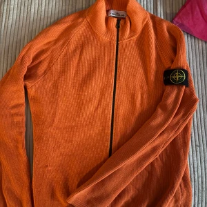Orange stickad kofta från Stone Island - Säljer en orange stickad kofta från Stone Island med hel dragkedja och klassisk logga på ärmen. Tröjan har ribbad struktur, hög krage och lång ärm. Perfekt för dig som vill sticka ut med en färgstark och snygg look. Köpt på NK i Stockholm för 8999 och den går inte att köpa längre.