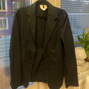 Tunn kavaj från Arket, storlek 48 - Säljer en kavaj från Arket i storlek 48. Kavajen är tillverkad i 100% bomull och har klassisk krage, två knappar framtill och tre utanpåliggande fickor. Perfekt för en stilren och enkel look.