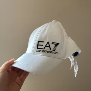 Vit EA7 Emporio Armani keps - Snygg vit keps från EA7 Emporio Armani med svart broderad logga framtill. Klassisk böjd skärm och justerbar baksida. Tillverkad i mjuk bomull som känns skön mot huvudet. Storlek One Size. Använd en gång, i nyskick. 