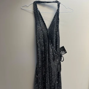 Glittrig svart långklänning - Supersnygg svart långklänning från Nelly med massor av glittriga paljetter. Djup v-ringning och omlott-design framtill. Perfekt för dig som vill sticka ut på festen! Klänningen har ärmlös halterneck och markerad midja.