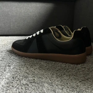 Svarta Maison Margiela sneakers - Maison Margiela sneakers i svart skinn och mocka med vita snören och beige insida. Snygg kontrast mellan material och färger, samt klassisk gummisula i brunt. Perfekt för dig som gillar stilrena och tidlösa sneakers med cool retrovibe.