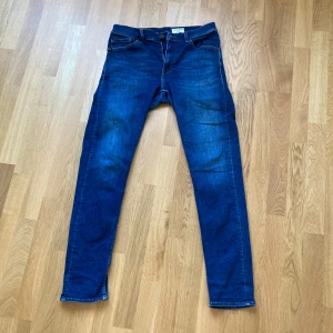 Blå jeans Tiger of sweden Evolve 32/32 - Snygga blå jeans från Tiger of Sweden i klassisk femficksmodell. Jeansen har en rak passform med normal midja och dragkedja. Materialet är slitstarkt denim i bomull och färgen är en djup blå med lätt slitningar framtill för en cool look.