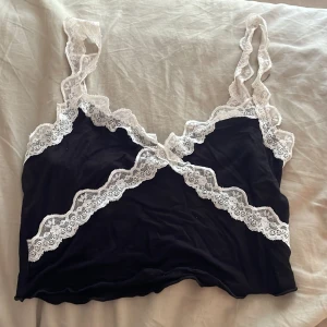 Svart spetslinnetopp med vita detaljer - Supersöt svart linnetopp med vita spetsdetaljer längs kanterna och axelbanden. Toppen har en croppad passform och tunna axelband i spets. Materialet är mjukt och stretchigt med 95% viskos och 5% spandex, vilket gör den riktigt bekväm.