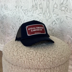 Marinblå keps John Hatter & Co.      Nyskick!! - Snygg marinblå truckerkeps från John Hatter & Co med mesh på sidorna och justerbar snapback. Framtill sitter en röd skylt med texten 'AFTER ALL TOMORROW IS ANOTHER DAY'. Perfekt statement-piece för din outfit. Helt ny med original lappar 