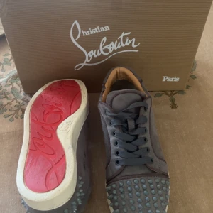Grå Christian Louboutin sneakers med nitar - Snygga grå sneakers från Christian Louboutin i mocka med ikoniska röda sulor och coola nitar på tån. Skorna har grå snören och beige insida. Passar 40/41,5
