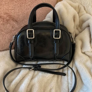 Svart handväska ZARA - Snygg svart handväska från Zara i glansigt skinn med två handtag och justerbar axelrem. Väskan har silverfärgade spännen framtill och dragkedja upptill. Den är i mycket fint skick💕