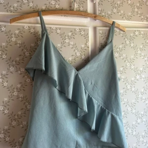 Ljusblå linnetopp med volang H&M XS - Supersöt ljusblå linnetopp från H&M i storlek XS. Toppen har smala axelband och en snygg volangdetalj som går diagonalt över framsidan. Perfekt för varma dagar och enkel att styla med jeans eller kjol.