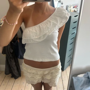 Vit oneshoulder topp med volang - Superfin vit oneshoulder topp med bred volangkant i broderad spets. Toppen är croppad och har en snygg asymmetrisk axel. Perfekt till sommaren och lätt att matcha med både jeans och kjol. Materialet känns mjukt och luftigt.