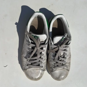 Adidas Stan Smith vita sneakers - Klassiska Adidas Stan Smith sneakers i vitt skinn med gröna detaljer på hälen och tungan. Skorna har rund tå, platt sula.