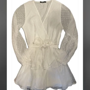 Vit volangklänning från Gina Tricot - Superfin vit kort volangklänning från Gina Tricot med knyt i midjan, långärmade transparenta ärmar med diskret prickigt mönster och v-ringning. Perfekt för student/kalas/avslutningar eller sommarens alla tillfällen! 