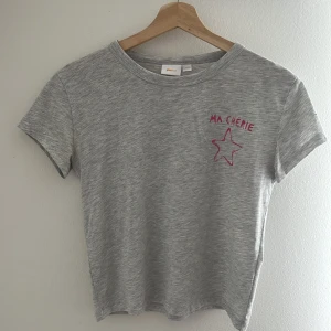 Grå t-shirt Gina Tricot med tryck - Gråmelerad t-shirt från Gina Tricot med korta ärmar och rund hals. På bröstet finns ett rosa tryck med texten 'MA CHÉRIE' och en stjärna. Skön basic i mjuk bomull, perfekt till jeans eller shorts.