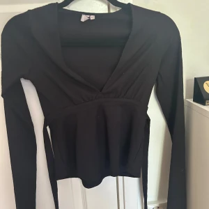 Svart långärmad v-ringad topp - Snygg svart långärmad topp med v-ringning och markerad midja. Modellen är croppad och har en lätt rynkad detalj under bysten. Perfekt att styla med jeans eller kjol för en trendig look.