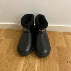 Svarta boots från Hugo med foder - Svarta boots från Hugo med mjukt, fluffigt foder och rund tå. Skorna har en blank finish och logga framtill. Perfekta för kyliga dagar när du vill hålla stilen och värmen.