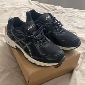 Asics sneakers - Svarta Asics sneakers med vita detaljer och robust sula. Ovandel i mesh och syntetläder, snörning framtill och klassisk Asics-logga på sidan. Perfekta för dig som gillar sportig stil och vill ha bekväma skor till vardagen.