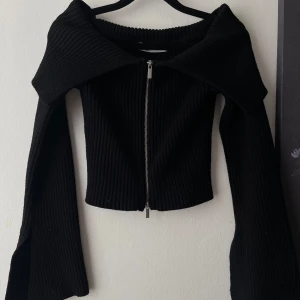 Svart off shoulder kofta med dragkedja - Säljer en svart ribbad off shoulder kofta med bred krage och dragkedja framtill. Tröjan har långa ärmar med lätt utsvängd form. 
