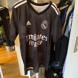 Real Madrid Benzema svart matchtröja - Snygg svart Real Madrid fotbollströja från Adidas med Benzema och nummer 9 på ryggen. Tröjan har vita detaljer, klubbmärke på bröstet och Emirates Fly Better-tryck framtill. Tillverkad i lätt och ventilerande material, perfekt för fotboll eller supporterliv.
