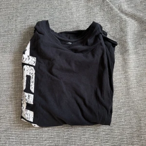 Svart t-shirt med vit text - Svart t-shirt i bomull med tryckt vit text längs sidan. Klassisk rund hals och korta ärmar, perfekt för en avslappnad stil. Enkel och snygg design som passar till det mesta.