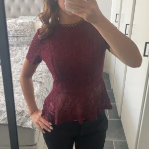 Vinröd spetsblus med volang - Supersnygg vinröd blus i spets med korta ärmar och volang nertill. Blusen har figurnära passform och transparenta detaljer som ger en extra cool touch. Perfekt för dig som vill sticka ut lite extra med en romantisk vibe.